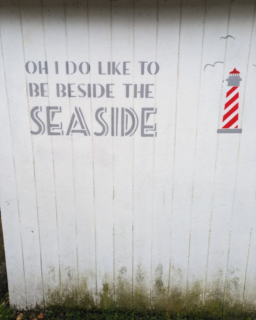 "Oh I do like to be beside the seaside" written on the back of a Bognor Regis beach hut next to a picture of a red and white striped lighthouse